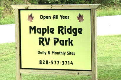 A wooden sign says "Open All Year - Maple Ridge RV Park - Daily & Monthly Sites - 828-577-3714"